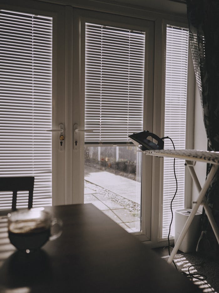 A sunlit indoor scene featuring an ironing board, blinds, and a tranquil atmosphere.